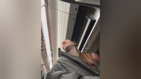 Media: Video of a young woman lying on a plane's overhead bin, wearing a gray hoodie, with her head resting on the bin's edge, eyes closed, and mouth slightly open.