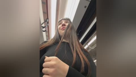 Media: Video of a young Asian woman with long, straight brown hair, wearing a black sweater, standing in a modern subway car with fluorescent lights and vertical wooden panels.