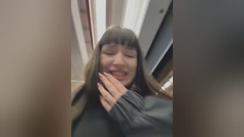 Media: Video of a young woman with long brown hair, smiling, wearing a black sweater, with blurred hands around her neck, in a dimly lit room with curtains and a white ceiling fan.