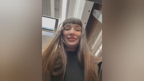 Media: Video of a young woman with long, straight, light brown hair styled in two braids. She has a light complexion, wearing a black top. The background features a modern office with vertical blinds and a computer monitor.