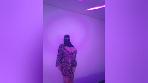 Media: A video of a person wearing a leopard-print dress with a long, black veil, standing in a dimly lit, minimalist room with purple lighting. The person's back is turned, creating a mysterious and enigmatic atmosphere.