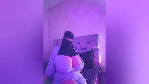 Media: Video of a curvy, light-skinned woman with large breasts wearing a nun's habit and mask, seated on a bed with a tufted headboard under purple lighting.