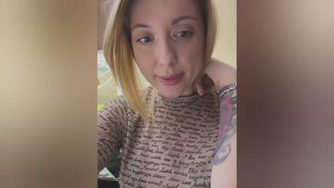 Media: Video of a middle-aged woman with light skin and shoulder-length blonde hair, wearing a patterned long-sleeve top. She has a small tattoo on her forearm and is smiling slightly, looking into the camera.