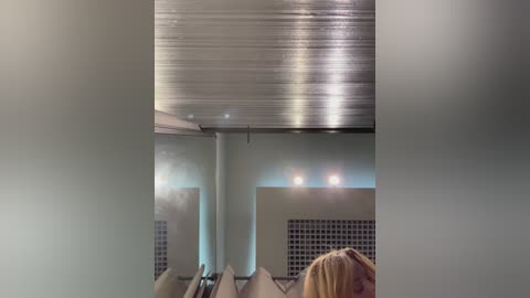 Media: Video of a modern spa interior featuring a metallic ceiling, soft lighting, and a reflection of a blonde woman with long hair, partially visible in the lower right.