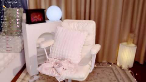 Media: Video of a plush white cat-shaped chair with a pink cushion, illuminated by a glowing orb and a candle, against beige curtains.