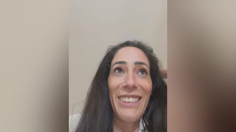 Media: Video of a middle-aged woman with long dark hair, light skin, and a wide smile, standing in a beige hallway. She has a playful, cheerful expression.