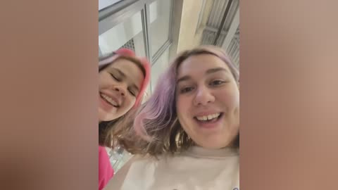 Media: Video of two young women, one with pink hair, smiling broadly, taken from a low angle; they are indoors near large windows.