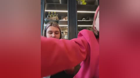 Media: Video of a young woman with light brown hair wearing a pink sweater, blurred in the foreground, standing in front of a dark, modern bookshelf with illuminated shelves.