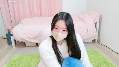 Media: Video of an East Asian woman with long black hair, wearing a beige face mask, white robe, and green carpet, in a pink-tiled bedroom.