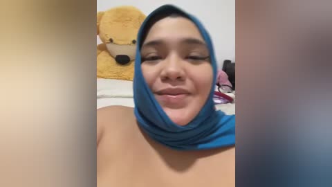 Media: A video of a woman with light skin, wearing a blue hijab, smiling softly, with a teddy bear and toys in the background.