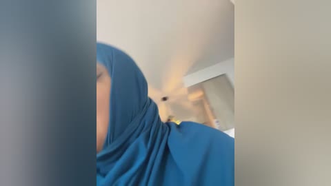 Media: A blurred video of a person wearing a blue hijab and a green shirt, standing indoors with a beige wall and a ceiling light in the background.