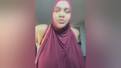 Media: A blurry video of a young woman with light brown skin, wearing a maroon hijab and a sleeveless top, indoors with a blurred background.