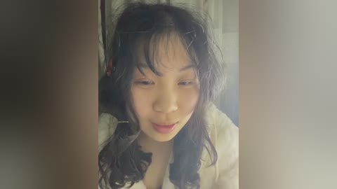 Media: A video of a young Asian woman with long black hair and a slight smile, wearing a white blouse with a black bow, standing indoors in a softly lit room.