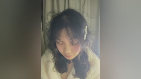 Media: Video of an Asian woman with dark curly hair, wearing a white robe, standing in a dimly lit room with a blurred curtain in the background.