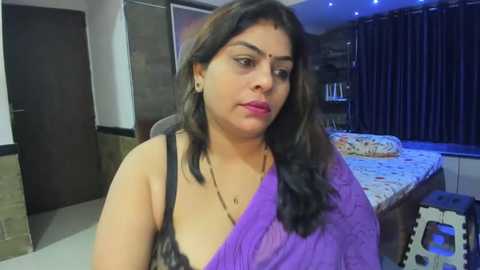 Media: Video of a South Asian woman with medium skin tone, wearing a purple sari and black top, sitting in a modern bedroom with dark curtains and a bed.