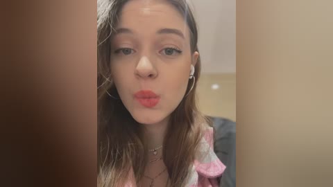 Media: Video of a young Caucasian woman with fair skin, straight brown hair, wearing a pink top, making a kissy face. Background is blurred, featuring beige walls.