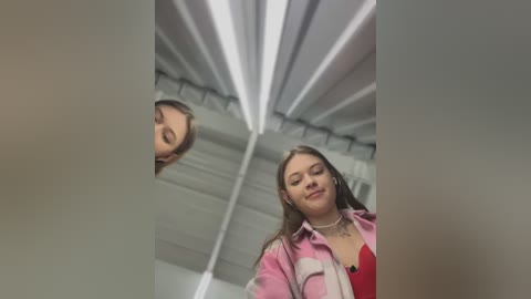 Media: Video of a young woman with long brown hair, wearing a pink and white striped shirt, standing in an industrial warehouse with exposed white beams.