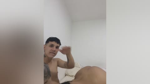 Media: Video of a shirtless, muscular man with short dark hair and a tattoo on his right shoulder, lying on a bed with white sheets, in a minimalistic, white room.