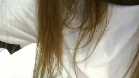 Media: Video of a person with long, straight brown hair partially covering their face, wearing a white shirt. The background is blurry, featuring white and black fabric.