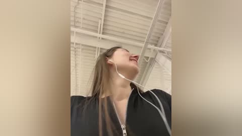 Media: Video of a woman with light skin, long brown hair, and a black jacket, wearing white earbuds, smiling and looking upward in an industrial setting with white ceilings and beams.