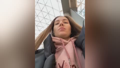 A video of a young woman with long, straight hair, wearing a pink hoodie and headphones, seated indoors under a modern glass ceiling with geometric patterns.