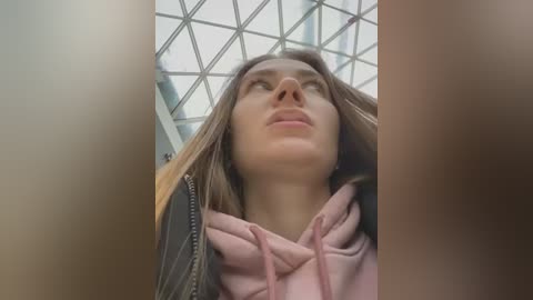 Media: Video of a young woman with long brown hair, wearing a pink hoodie, looking upward with a contemplative expression. The background features a modern, grid-patterned glass ceiling.