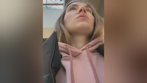 Media: A video shows a young woman with straight, shoulder-length light brown hair, wearing a pink hoodie, with a relaxed expression, looking upwards, taken from a low angle.