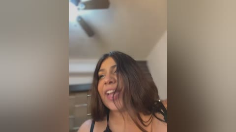 Media: Video of a smiling Latina woman with long dark hair, wearing a black bra, in a dimly lit room with beige walls and a ceiling fan.