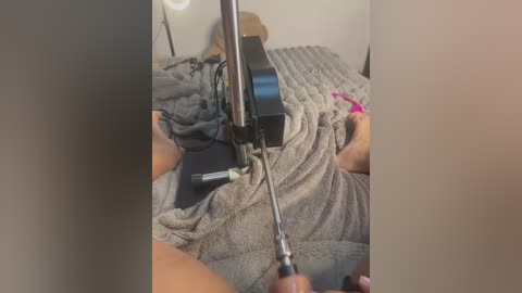 Media: A video shows a person with light skin using a screwdriver to adjust a black box on a bed with gray blankets. The background is a neutral-toned room.