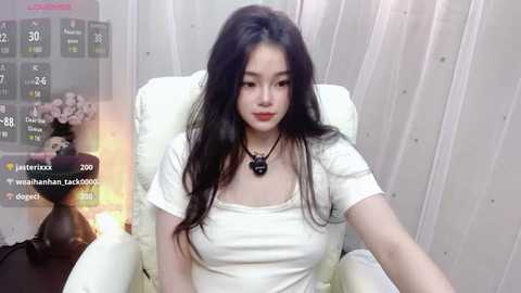 Media: A video of an East Asian woman with long black hair, fair skin, and a slender physique, sitting in a white chair. She wears a white top and a black necklace. Background includes a calendar and a partially visible person.