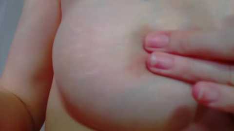 Media: A close-up video of a light-skinned woman's bare breast, with her hand gently cupping it. Her fingernails are unpolished. The background is blurred, focusing attention on the breast and hand.