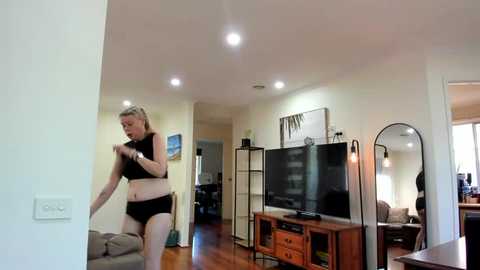 Media: A video of a woman with blonde hair in black workout clothes adjusting a lamp in a modern living room with wooden floors, a flat-screen TV, and a mirrored closet.