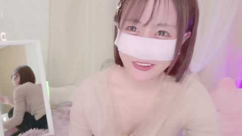 A video of an Asian woman with straight, shoulder-length brown hair, wearing a white mask and a beige top, smiling, with a mirror reflecting her back.