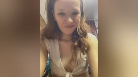 Media: Video of a young Caucasian woman with wavy brown hair, wearing a low-cut, white crop top, smiling slightly, indoors.