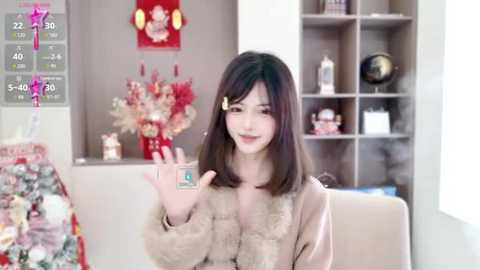 Media: Video of an Asian woman with shoulder-length brown hair, wearing a beige fur coat, holding a small package. Background features a modern, minimalist room with a bookshelf and floral arrangement.