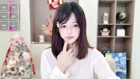 Video of a young Asian woman with straight black hair and white hair accessory, wearing a white sweater, standing in a modern, organized room with a Christmas tree and shelves filled with decorative items.