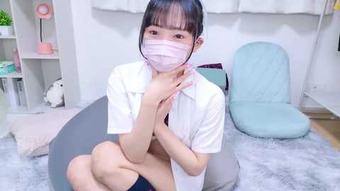 Video of an East Asian woman with light skin, straight black hair, wearing a white shirt, pink face mask, and no pants, sitting on a grey bean bag chair in a minimalist, white room with a light blue chair.