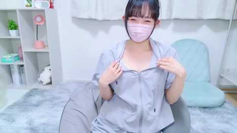 Video of an Asian woman in a light gray hospital gown, sitting on a light blue chair, wearing a pink mask, in a bright, modern room with white walls and a gray rug.