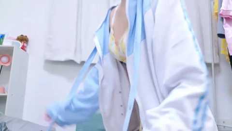 Video of a woman in a white lab coat, partially unbuttoned, revealing a yellow bra, holding blue gloves, in a medical setting with white walls and medical supplies.