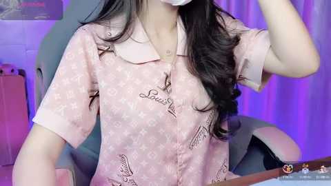 Media: Video of an Asian woman with long black hair, wearing a light pink, short-sleeved, star-patterned Louis Vuitton pajama shirt, sitting in a grey gaming chair, with purple curtains in the background.