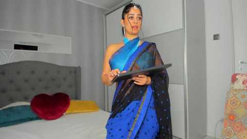 Media: Video of a South Asian woman in a blue sari, holding a tray, standing in a modern, minimalist bedroom with a tufted headboard, red heart pillow, and white walls.