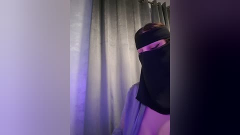 Media: Video of a person wearing a black blindfold with a pink stripe, standing in a dimly lit room with grey curtains.