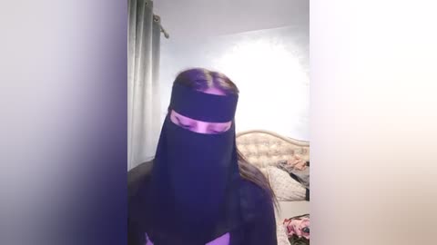 Media: A video of a person wearing a black burqa with a pink bandana, standing in a dimly lit room with a bed, pink sheets, and a beige headboard.