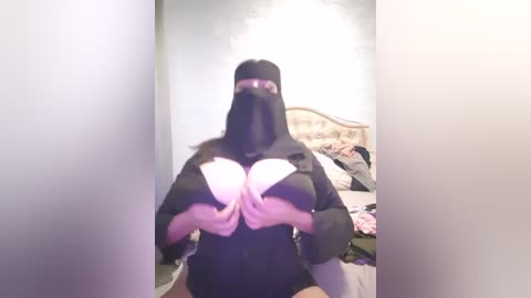 Media: Video of a person in a black nun costume, holding white bra cups in front of the face, standing in a messy bedroom with a bed and scattered clothes.