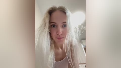 Media: A video of a young Caucasian woman with long, platinum blonde hair, light skin, and blue eyes, wearing a white tank top, taken indoors with a blurred background.