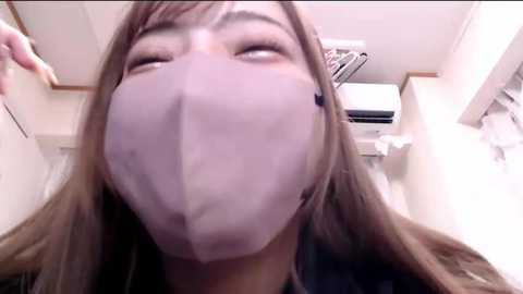 Media: Video of a young woman with light brown hair, wearing a light purple face mask, taken from a low angle, showing her smiling face and upper body in a white room with an air conditioner.