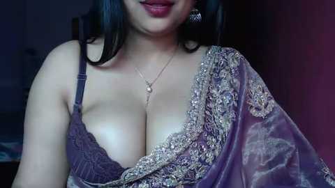 Media: Video of a South Asian woman with medium skin tone, wearing a purple, embroidered sari with a lace bra, revealing ample cleavage. She has dark hair and wears a gold necklace and earrings.