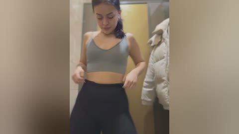 Media: A video of a young woman with light skin and straight dark hair, wearing a grey sports bra and black leggings, adjusting her leggings in a narrow hallway.