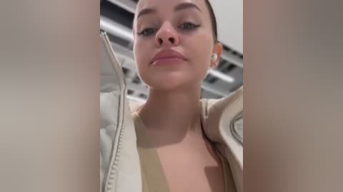 Media: Video of a young woman with light skin and dark hair, wearing a beige jacket, standing indoors with metallic ceiling panels in the background.