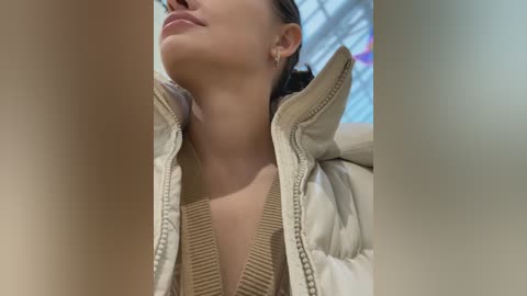 Media: Video of a young woman with light skin and dark hair, wearing a beige puffer jacket and a beige ribbed sweater, gazing upward in a bright, modern indoor setting.